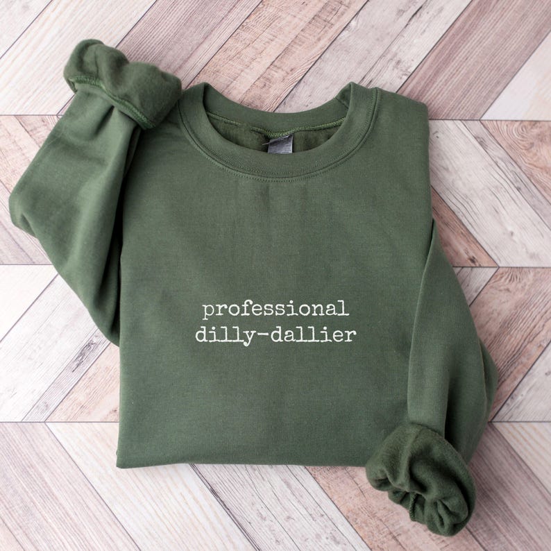 May include: An olive green sweatshirt with the phrase "professional dilly-dallier" printed in white. The sweatshirt is folded on a light brown wooden surface.