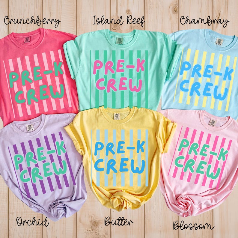 May include: Six brightly coloured t-shirts with the text "PRE-K CREW" in a striped design. The colours are pink, light green, light blue, lavender, yellow, and light pink. The shirts are displayed on a light wood surface.