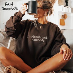 May include: A dark chocolate brown crewneck sweatshirt featuring the text professional homebody embroidered in a light beige thread across the chest. The long-sleeved garment has a relaxed fit and is worn with a gold necklace and rings.