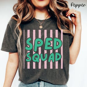 May include: A charcoal gray short-sleeve t-shirt featuring a graphic design of vertical light pink stripes. Overlaid on the stripes is the text "SPED SQUAD" in a bold, rounded, bright green font. The shirt has a crew neckline and a relaxed fit.