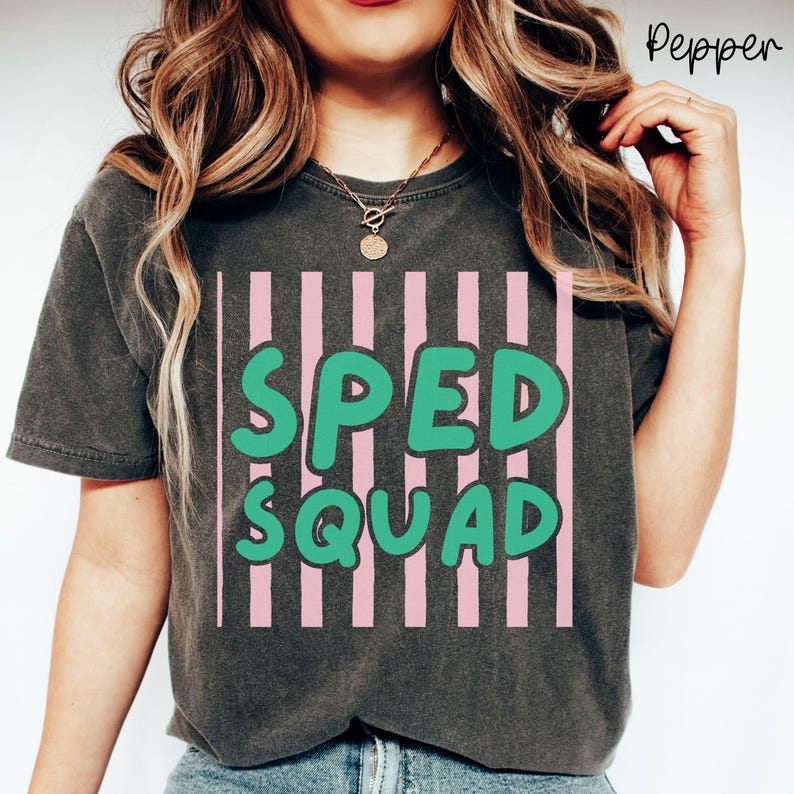 May include: A charcoal gray short-sleeve t-shirt featuring a graphic design of vertical light pink stripes. Overlaid on the stripes is the text "SPED SQUAD" in a bold, rounded, bright green font. The shirt has a crew neckline and a relaxed fit.