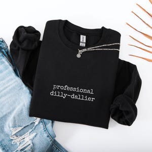 May include: Black crewneck sweatshirt with the text "professional dilly-dallier" in white. A silver necklace with a circular pendant is draped across the sweatshirt. Faded blue jeans and a black sleeve are visible.