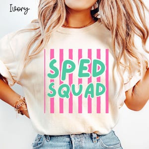 May include: An ivory short-sleeved t-shirt featuring a graphic design with vertical pink stripes and the text "SPED SQUAD" in large, bold, light green letters. The shirt has a crew neckline and a relaxed fit, with the word "Ivory" printed in the corner.