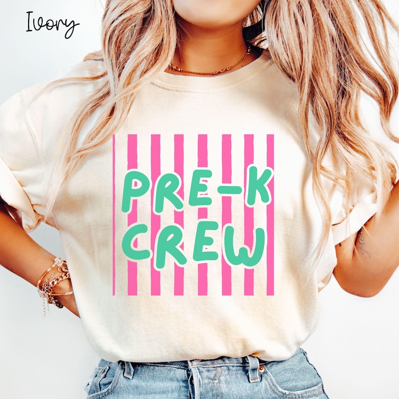 May include: Ivory-coloured sweatshirt featuring the text "PRE-K CREW" in green, set against a pink striped background. A casual style, suitable for everyday wear.