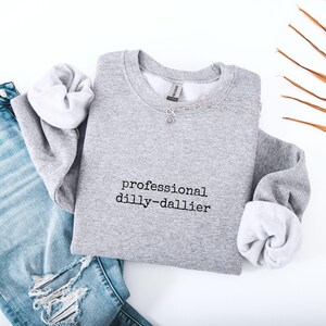 May include: A heather gray sweatshirt with the text "professional dilly-dallier" in black. The sweatshirt has a crew neck and long sleeves with white cuffs. A silver necklace with a pendant is draped across the neckline. Light blue jeans are visible.