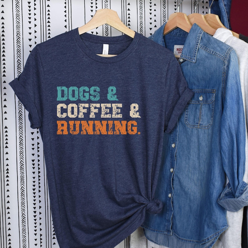 May include: Navy blue t-shirt with the text "DOGS & COFFEE & RUNNING" in teal, cream, and orange. The shirt is hanging on a wooden hanger with other shirts in the background.