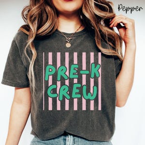 May include: Dark grey t-shirt featuring the text "PRE-K CREW" in green bubble letters, set against pink vertical stripes. The t-shirt is a casual style.