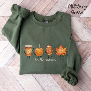 May include: A military green crewneck sweatshirt featuring a graphic print of a coffee cup, a caramel apple, a football, and an orange maple leaf, each with a smiling face and stick arms and legs, with the text 'tis the season' below.