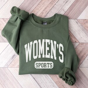 May include: An olive green sweatshirt with the text "WOMEN'S SPORTS" in white block letters. The sweatshirt is folded on a light brown and white wooden surface. The sleeves are cuffed.