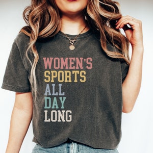 May include: A charcoal gray short-sleeve t-shirt featuring the text "WOMEN'S SPORTS ALL DAY LONG" printed in stacked lines. The word "WOMEN'S" is in pink, "SPORTS" is in yellow, "ALL" is in light blue, "DAY" is in teal, and "LONG" is in off-white.