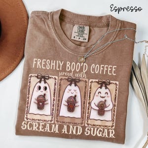 May include: An espresso-colored t-shirt featuring a graphic of three ghosts holding coffee drinks. The text reads "FRESHLY BOO'D COFFEE served with SCREAM AND SUGAR". The shirt has a crew neckline and a Comfort Colors brand label at the inner collar.