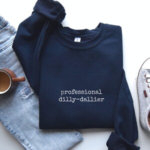 May include: Navy blue sweatshirt with the text "professional dilly-dallier" in white. The sweatshirt is folded, with a pair of blue jeans, a coffee mug, and a sneaker visible in the background.