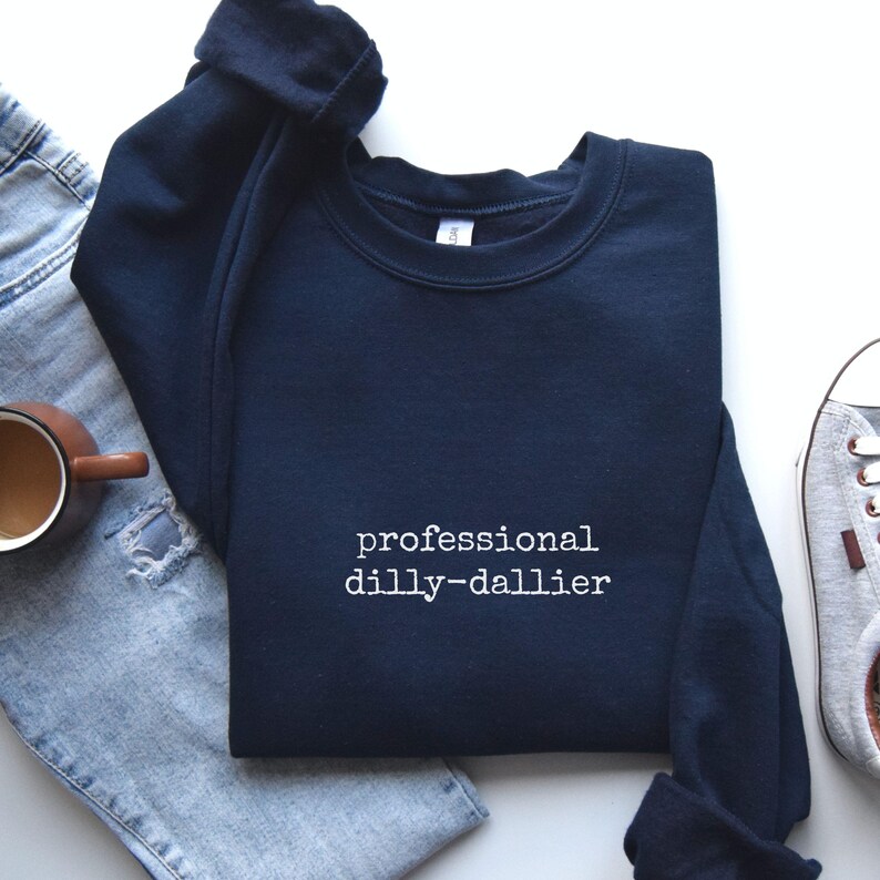 May include: Navy blue sweatshirt with the text "professional dilly-dallier" in white. The sweatshirt is folded, with a pair of blue jeans, a coffee mug, and a sneaker visible in the background.