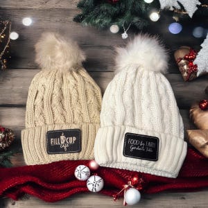 May include: Two white knit winter hats with faux fur pom poms. One hat has a brown leather patch with the text "Fill 'er Up Cafe" and the other has a brown leather patch with the text "Food by Faith Garden to Table".