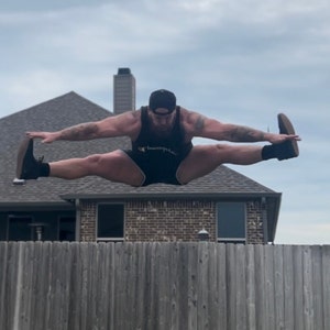 May include: A man wearing a black tank top with the word "Champion" on it, black shorts, and a black baseball cap is jumping in the air with his legs spread wide. He is wearing black trainers. The background is a brick house and a wooden fence.