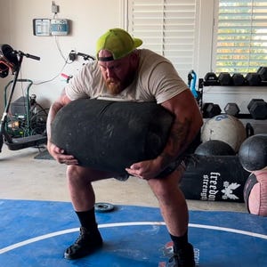 Może przedstawiać: Mężczyzna w zielonej czapce bejsbolowej podnosi duży czarny ciężar w garażowej siłowni. Ciężar znajduje się na niebieskiej macie do zapasów z nadrukiem "Freedom Strength" i "Official Mat".