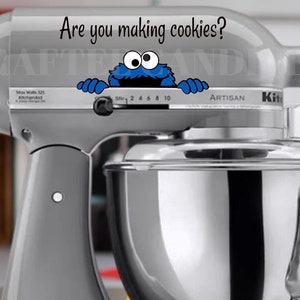 May include: A gray stand mixer with a silver bowl. A blue cookie monster character is peeking over the top of the mixer with the text "Are you making cookies?" above it.