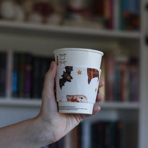 Könnte beinhalten: Ein weißer Papier-Kaffeebecher mit einer Stoffmanschette im Halloween-Design. Die Manschette zeigt Illustrationen von braunen Fledermäusen, orangefarbenen Hexenhüten, kleinen gelben Sternen und einem Geist, der einen Kürbis auf weißem Grund hält.