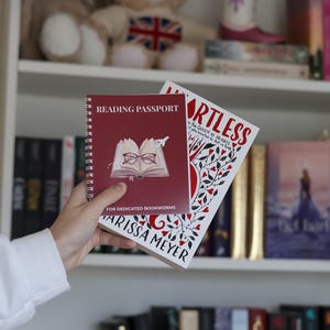 Op de afbeelding: Een bordeauxrood spiraalgebonden Reading Passport notitieboek met een illustratie van een open boek met een bril en een klein vliegtuigje, vastgehouden naast een wit boek getiteld Heartless van Marissa Meyer met rode hartjes en ranken.