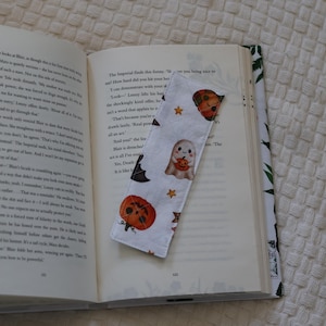 Op de afbeelding: Een rechthoekige stoffen boekenlegger met een witte achtergrond en een herhalend patroon van oranje pompoenen, lachende spookjes met pompoenen, kleine oranje sterretjes en zwarte vleermuisvleugels, liggend op de pagina's van een boek.