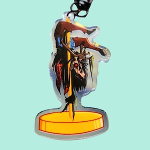 Pode incluir: Um chaveiro de acrílico colorido com um veado de desenho animado com um olho vermelho, vestindo uma camisa azul e calças vermelhas, pendurado de cabeça para baixo em um poste dourado. O veado está rodeado de folhagem verde.