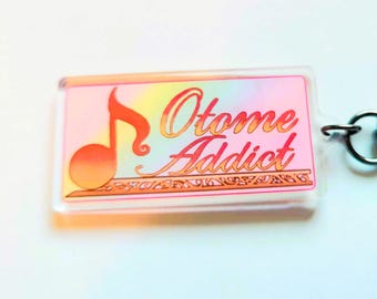 May include: A clear acrylic keychain with a pink and orange ombre background. The keychain features a musical note and the text "Otome Addict" in pink.
