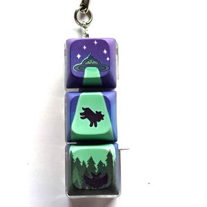 May include: A keychain with three keycaps featuring different designs. The top keycap is purple with a green UFO and stars. The middle keycap is green with a black silhouette of a creature. The bottom keycap is green with a black silhouette of a bat and trees.
