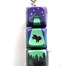 May include: A keychain with three keycaps featuring different designs. The top keycap is purple with a green UFO and stars. The middle keycap is green with a black silhouette of a creature. The bottom keycap is green with a black silhouette of a bat and trees.