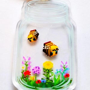 May include: A clear glass jar keychain with a yellow and black bee, a sunflower, and other flowers inside. The jar has a gold metal clasp.