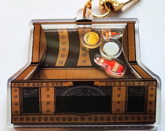 May include: A gold-colored metal keychain shaped like a treasure chest with a clear plastic front. Inside the chest are small, colorful, plastic trinkets, including a gold coin, a red bottle, and a white candy cane.