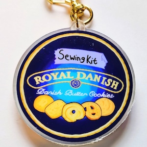 Peut inclure: Un porte-clés rond en acrylique transparent avec un design bleu et or. Le design présente le texte "Royal Danish Danish Butter Cookies" et une étiquette qui dit "Sewing Kit".