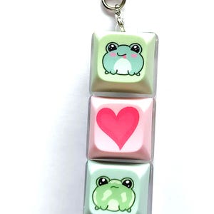 May include: A keychain with three keycaps. The top keycap is light green with a cartoon frog. The middle keycap is pink with a red heart. The bottom keycap is light green with a cartoon frog.