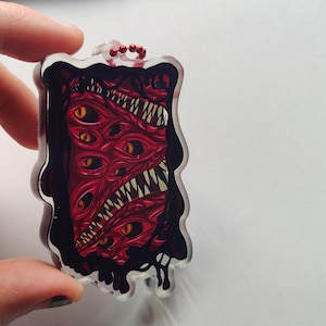 May include: A rectangular keychain featuring a red, multi-eyed monster design with sharp teeth and a black border. The keychain has a clear plastic loop with a small red bead chain.