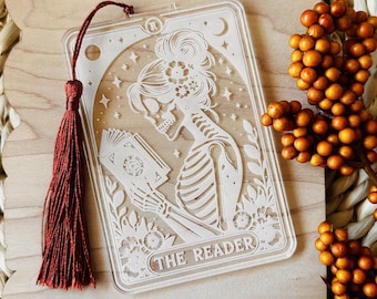May include: A white acrylic bookmark featuring a skeleton reading a book with the text "The Reader". The bookmark has a red tassel.