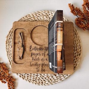 May include: A wooden tray with a quote about reading, "Between the pages of a book is a lovely place to be". The tray has a built-in cup holder and a slot for glasses. A book titled "Layla" by Colleen Hoover is propped up on the tray.