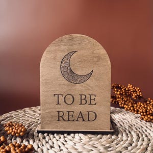 May include: Wooden arched book stand with a crescent moon design and the words "TO BE READ" engraved on the front. The stand is light brown and sits on a woven mat with decorative berries.
