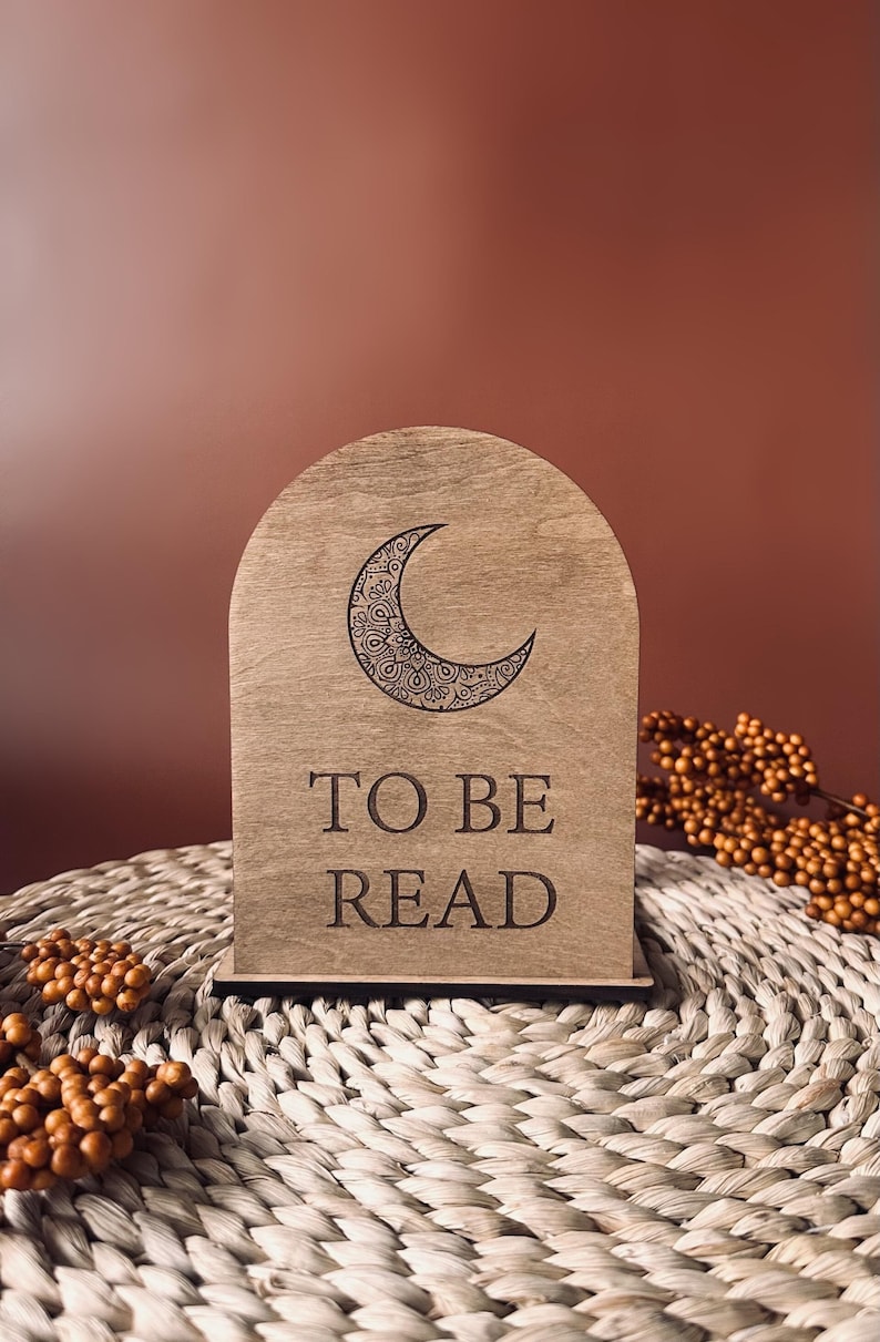 May include: Wooden arched book stand with a crescent moon design and the words "TO BE READ" engraved on the front. The stand is light brown and sits on a woven mat with decorative berries.