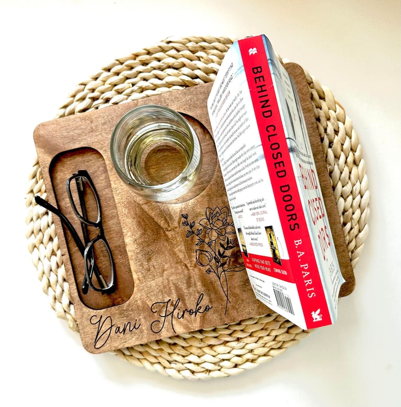 May include: A personalized wooden tray with a glass, glasses, and a book titled "Behind Closed Doors" by B.A. Paris. The tray has a floral design and the name "Dani Hiroko" engraved on it.