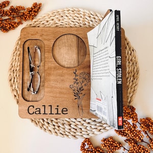 May include: A personalized wooden tray with a cutout for a cup, a slot for glasses, and a book holder. The tray is engraved with the name "Callie" and a floral design. The book is titled "Girl Stolen" by April Henry.