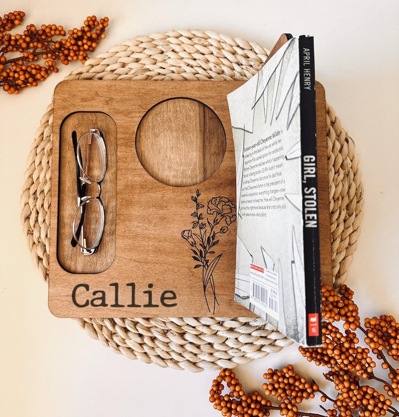May include: A personalized wooden tray with a cutout for a cup, a slot for glasses, and a book holder. The tray is engraved with the name "Callie" and a floral design. The book is titled "Girl Stolen" by April Henry.