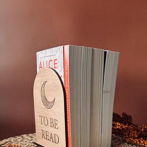 May include: A wooden bookend with a crescent moon design and the words "TO BE READ" is holding a stack of books. The bookend is light brown and the books have white pages. The background is a solid brown color.