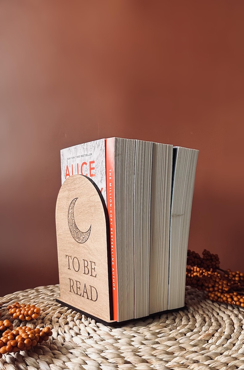May include: A wooden bookend with a crescent moon design and the words "TO BE READ" is holding a stack of books. The bookend is light brown and the books have white pages. The background is a solid brown color.