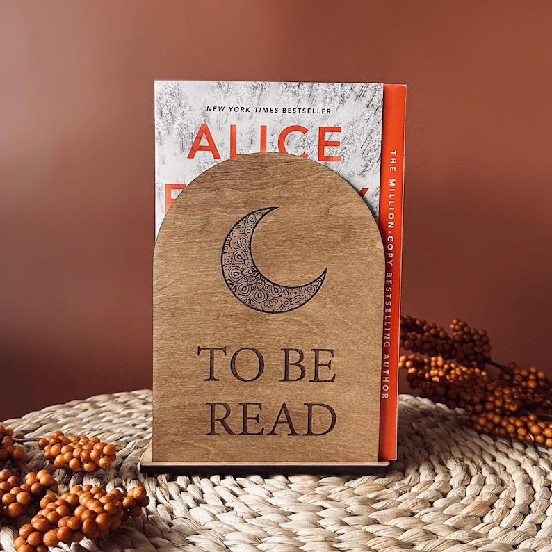 May include: Wooden bookend with a crescent moon design and the words "TO BE READ" engraved on the front. The bookend holds a book with a white and red cover. The bookend is made of light brown wood.