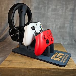 Puede incluir: Un soporte de madera con el texto "Gamer Dad Like a Normal Dad Only Much Cooler" sostiene un auricular inalámbrico negro, un mando Xbox blanco y verde y un mando Xbox rojo.