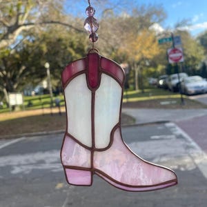 May include: A stained glass cowboy boot suncatcher in shades of pink, white, and burgundy. The boot is suspended by a thin wire with three purple beads. The suncatcher is designed to hang in a window.