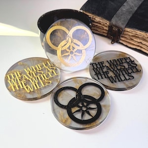 May include: Four round coasters with a marbled effect and gold or black designs. The coasters feature the text "THE WHEEL WEAVES AS THE WHEEL WILLS" and a stylized wheel symbol. A dark book and container are in the background.