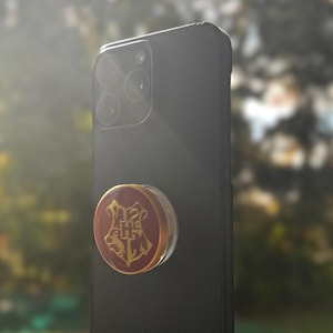 May include: A black smartphone with a circular phone grip attached to the back. The grip features a maroon circular face with a gold-colored crest design in the center, depicting a shield with various animal figures and the letters H and H.