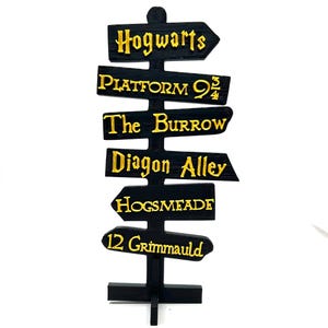 May include: Black wooden signpost with gold lettering pointing to various locations from the Harry Potter universe, including Hogwarts, Platform 9 3/4, The Burrow, Diagon Alley, Hogsmeade, and 12 Grimmauld Place.