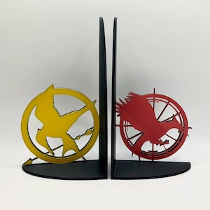 May include: A pair of black bookends with a Mockingjay symbol on each. The left bookend features a yellow Mockingjay with a black arrow, while the right bookend features a red Mockingjay with a black arrow.
