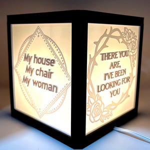 May include: A black wooden box with a white light inside. The box has two sides with text. One side says "My house, My chair, My woman." The other side says "There you are, I've been looking for you."